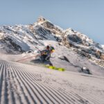 Ein Skifahrer in Winterausrüstung fährt eine frisch präparierte Piste hinunter, während im Hintergrund ein schneebedeckter Berg zu sehen ist.