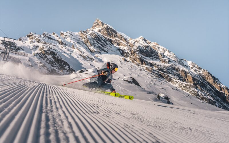 Ein Skifahrer in Winterausrüstung fährt eine frisch präparierte Piste hinunter, während im Hintergrund ein schneebedeckter Berg zu sehen ist.
