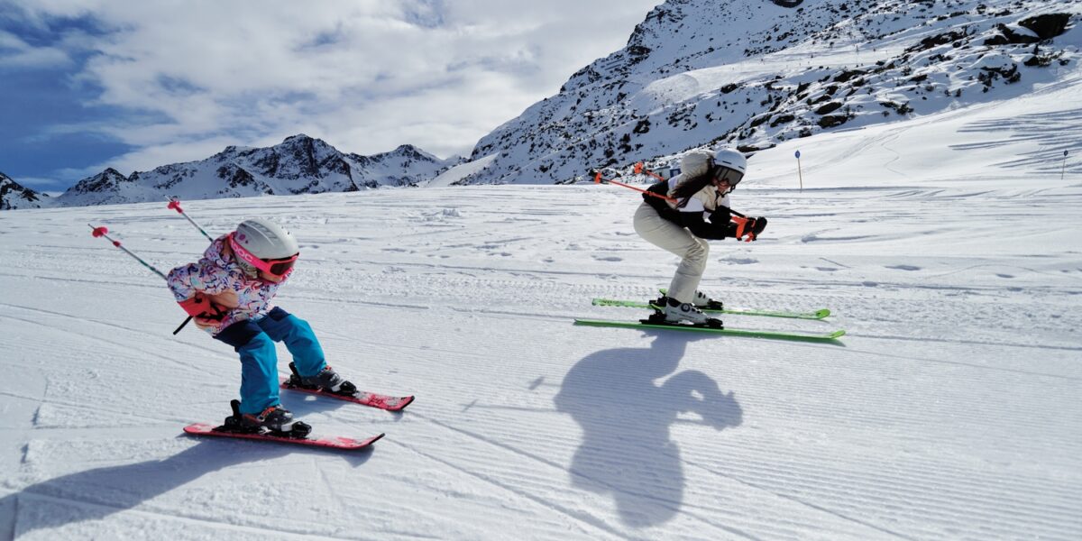 Zwei Skifahrer, ein Erwachsener und ein Kind, fahren auf einer verschneiten Bergpiste bei teilweise bewölktem Himmel bergab, mit felsigen Gipfeln im Hintergrund.
