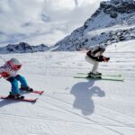 Zwei Skifahrer, ein Erwachsener und ein Kind, fahren auf einer verschneiten Bergpiste bei teilweise bewölktem Himmel bergab, mit felsigen Gipfeln im Hintergrund.