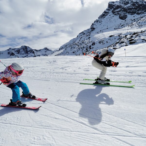 Zwei Skifahrer, ein Erwachsener und ein Kind, fahren auf einer verschneiten Bergpiste bei teilweise bewölktem Himmel bergab, mit felsigen Gipfeln im Hintergrund.