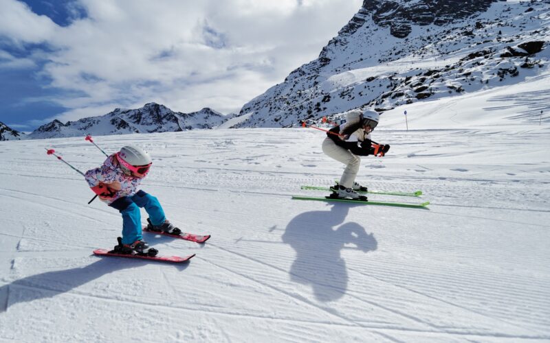 Zwei Skifahrer, ein Erwachsener und ein Kind, fahren auf einer verschneiten Bergpiste bei teilweise bewölktem Himmel bergab, mit felsigen Gipfeln im Hintergrund.