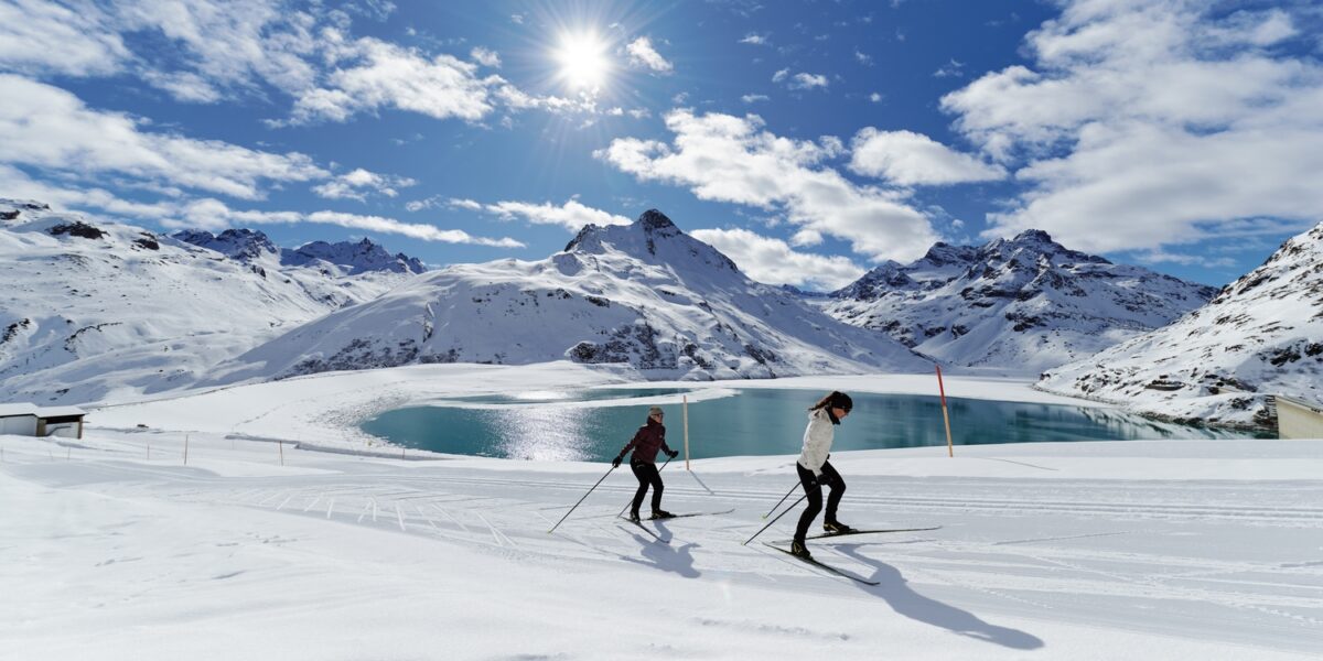 Zwei Langläufer auf einer schneebedeckten Loipe neben einem Bergsee, mit schneebedeckten Gipfeln und einer hellen Sonne im Hintergrund.