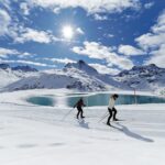 Zwei Langläufer auf einer schneebedeckten Loipe neben einem Bergsee, mit schneebedeckten Gipfeln und einer hellen Sonne im Hintergrund.