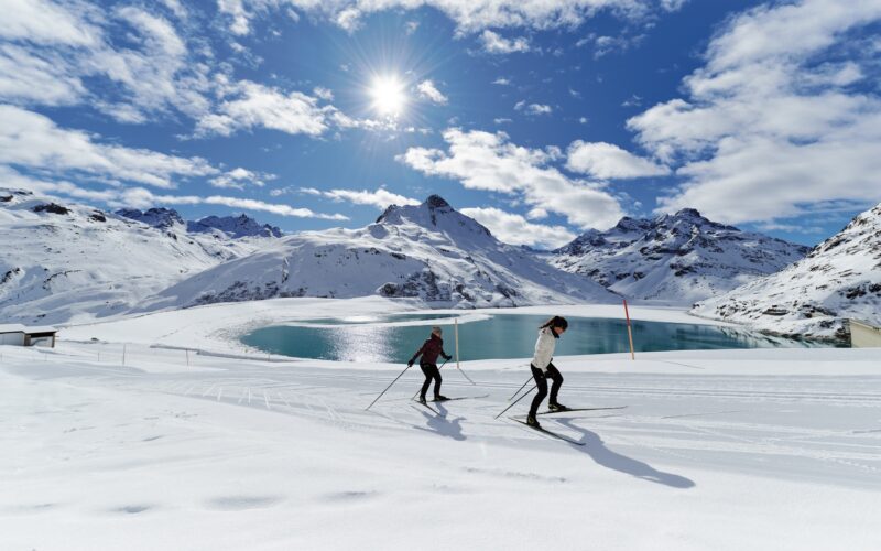 Zwei Langläufer auf einer schneebedeckten Loipe neben einem Bergsee, mit schneebedeckten Gipfeln und einer hellen Sonne im Hintergrund.