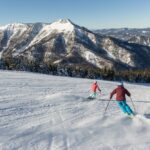 Zwei Skifahrer fahren einen verschneiten Hang hinunter, mit bewaldeten Bergen und einem klaren blauen Himmel im Hintergrund.