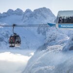 Eine Gondelbahn mit der Aufschrift "Panorama Jet Zwieselalm" befördert die Fahrgäste über eine verschneite Berglandschaft mit schroffen Gipfeln im Hintergrund.