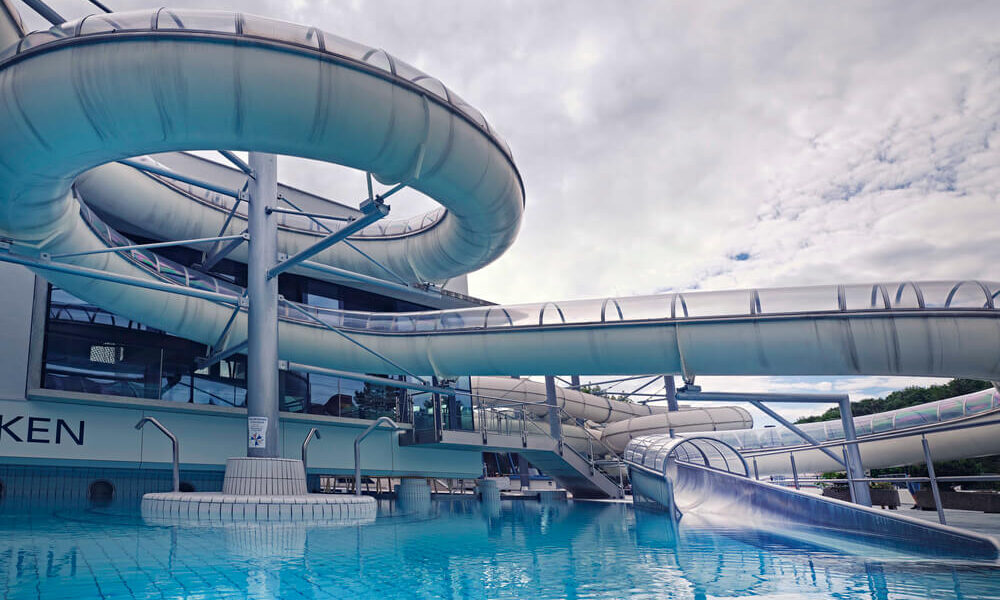 Große, geschlossene Wasserrutschen wölben sich über einem blau gekachelten Schwimmbecken in einem Indoor-Wasserpark unter einem bewölkten Himmel.