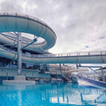 Große, geschlossene Wasserrutschen wölben sich über einem blau gekachelten Schwimmbecken in einem Indoor-Wasserpark unter einem bewölkten Himmel.