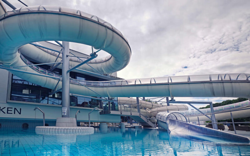 Große, geschlossene Wasserrutschen wölben sich über einem blau gekachelten Schwimmbecken in einem Indoor-Wasserpark unter einem bewölkten Himmel.