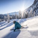 Ein Skifahrer in blauer Kleidung macht eine scharfe Kurve auf einer präparierten, verschneiten Piste, die von Kiefern und Bergen umgeben ist, bei klarem Himmel und Sonnenschein.