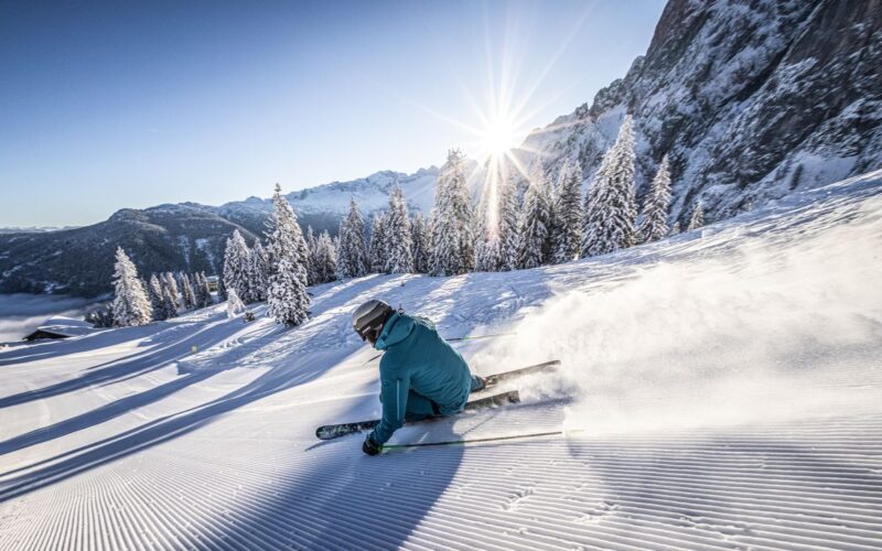 Ein Skifahrer in blauer Kleidung macht eine scharfe Kurve auf einer präparierten, verschneiten Piste, die von Kiefern und Bergen umgeben ist, bei klarem Himmel und Sonnenschein.