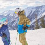 Zwei Personen in Skiausrüstung stehen auf einem verschneiten Berghang und halten Skistöcke. Im Hintergrund sind schneebedeckte Berge und ein bewölkter Himmel zu sehen.