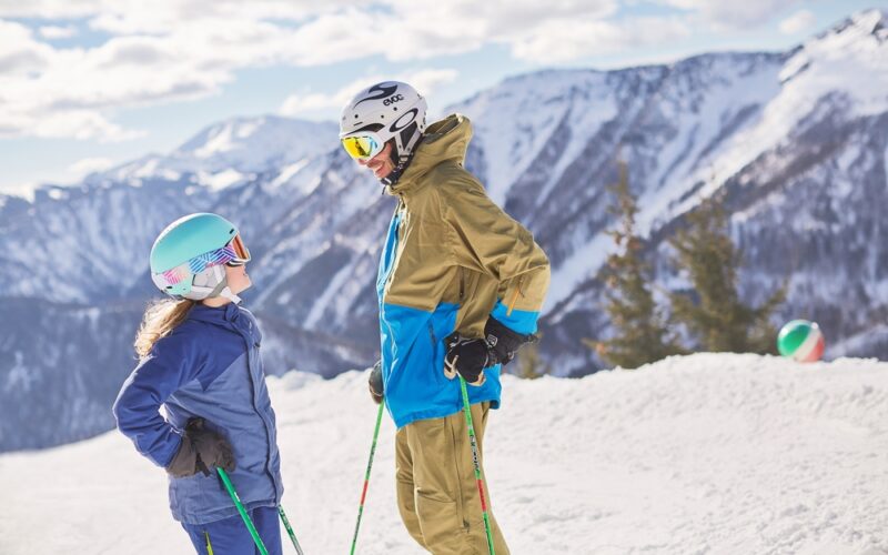 Zwei Personen in Skiausrüstung stehen auf einem verschneiten Berghang und halten Skistöcke. Im Hintergrund sind schneebedeckte Berge und ein bewölkter Himmel zu sehen.