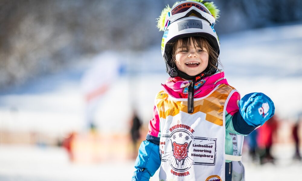 Ein kleines Kind in bunter Skikleidung und mit Helm lächelt auf einer verschneiten Piste und trägt eine Skischulweste mit einem Tiger-Logo.