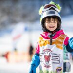 Ein kleines Kind in bunter Skikleidung und mit Helm lächelt auf einer verschneiten Piste und trägt eine Skischulweste mit einem Tiger-Logo.