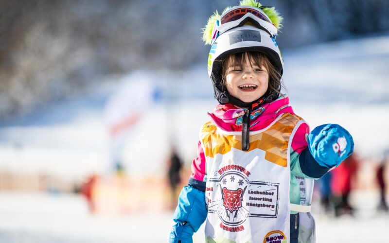 Ein kleines Kind in bunter Skikleidung und mit Helm lächelt auf einer verschneiten Piste und trägt eine Skischulweste mit einem Tiger-Logo.