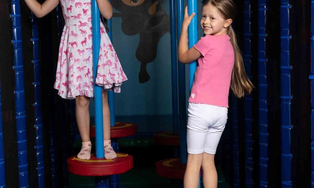 Zwei junge Mädchen stehen auf roten, kreisförmigen Plattformen in einem Indoor-Spielbereich und halten sich an senkrechten blauen Stangen fest, während hinter ihnen eine Ninja-Illustration an der Wand hängt.