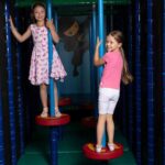 Zwei junge Mädchen stehen auf roten, kreisförmigen Plattformen in einem Indoor-Spielbereich und halten sich an senkrechten blauen Stangen fest, während hinter ihnen eine Ninja-Illustration an der Wand hängt.