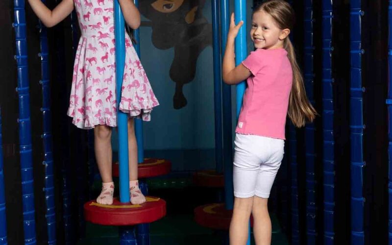 Zwei junge Mädchen stehen auf roten, kreisförmigen Plattformen in einem Indoor-Spielbereich und halten sich an senkrechten blauen Stangen fest, während hinter ihnen eine Ninja-Illustration an der Wand hängt.