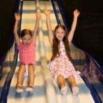 Zwei junge Mädchen rutschen mit erhobenen Händen eine zweispurige Indoor-Rutsche hinunter. Die eine trägt ein rosa T-Shirt und Shorts, die andere ein weißes Kleid mit rosa Motiven.