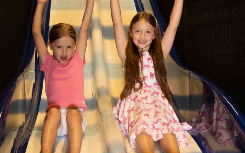 Zwei junge Mädchen rutschen mit erhobenen Händen eine zweispurige Indoor-Rutsche hinunter. Die eine trägt ein rosa T-Shirt und Shorts, die andere ein weißes Kleid mit rosa Motiven.