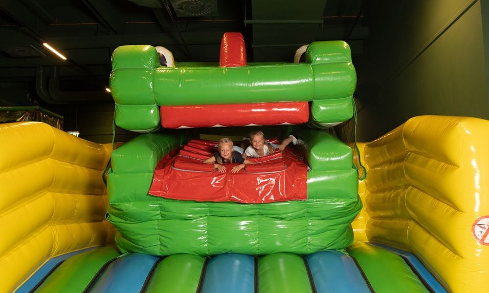 Zwei Kinder spielen in einem grün-gelben aufblasbaren Gebilde in Form eines Tieres mit offenem Maul.