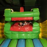 Zwei Kinder spielen in einem grün-gelben aufblasbaren Gebilde in Form eines Tieres mit offenem Maul.