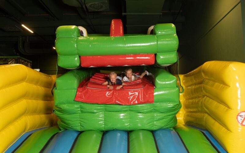 Zwei Kinder spielen in einem grün-gelben aufblasbaren Gebilde in Form eines Tieres mit offenem Maul.