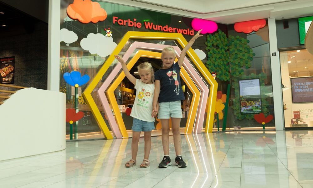 Zwei kleine Kinder stehen händchenhaltend und lächelnd vor einem bunten Schaufenster mit der Aufschrift "Farbic Wunderwald" in einem Einkaufszentrum.
