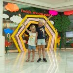 Zwei kleine Kinder stehen händchenhaltend und lächelnd vor einem bunten Schaufenster mit der Aufschrift "Farbic Wunderwald" in einem Einkaufszentrum.