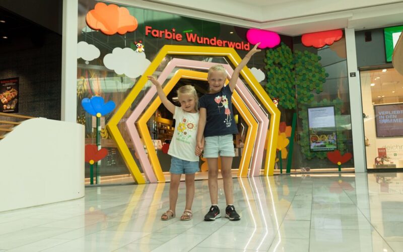 Zwei kleine Kinder stehen händchenhaltend und lächelnd vor einem bunten Schaufenster mit der Aufschrift "Farbic Wunderwald" in einem Einkaufszentrum.