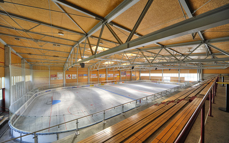 Sporthalle mit leeren Tribünenplätzen mit Blick auf eine mit roten und blauen Linien markierte Rollhockeybahn, umgeben von Sicherheitstafeln und Netzen.