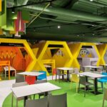 Ein moderner Indoor-Spielbereich mit geometrischen gelben Spielgeräten, buntem Deckendekor und Tischen mit grauen Stühlen im Vordergrund.