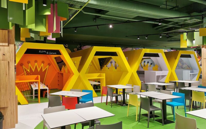 Ein moderner Indoor-Spielbereich mit geometrischen gelben Spielgeräten, buntem Deckendekor und Tischen mit grauen Stühlen im Vordergrund.