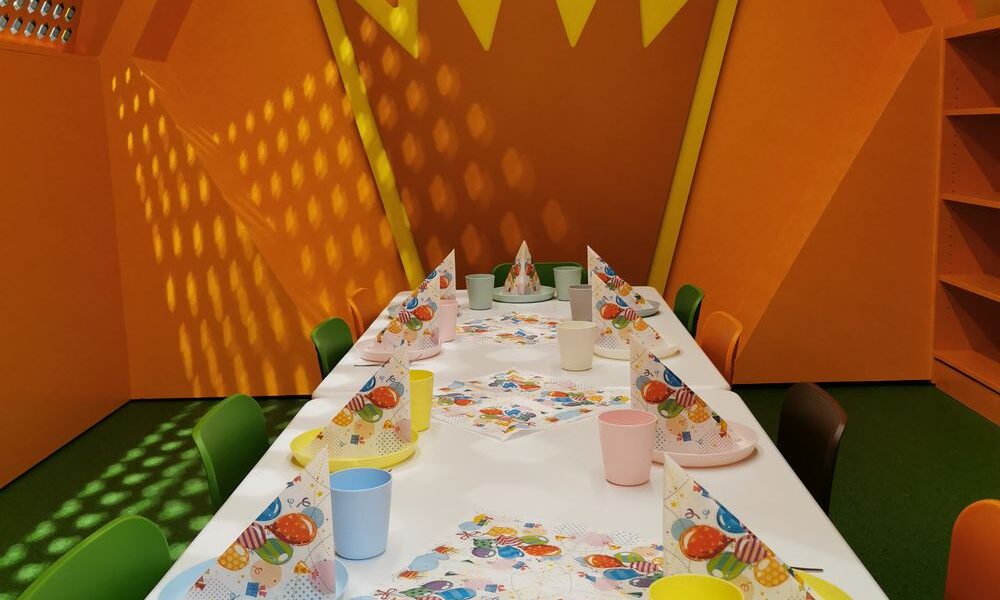 In einem Raum mit orangefarbenen Wänden und einer großen gelben Kronendekoration an der Rückwand ist ein bunter Partytisch mit Papierhüten, Bechern und Servietten gedeckt.