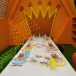 In einem Raum mit orangefarbenen Wänden und einer großen gelben Kronendekoration an der Rückwand ist ein bunter Partytisch mit Papierhüten, Bechern und Servietten gedeckt.