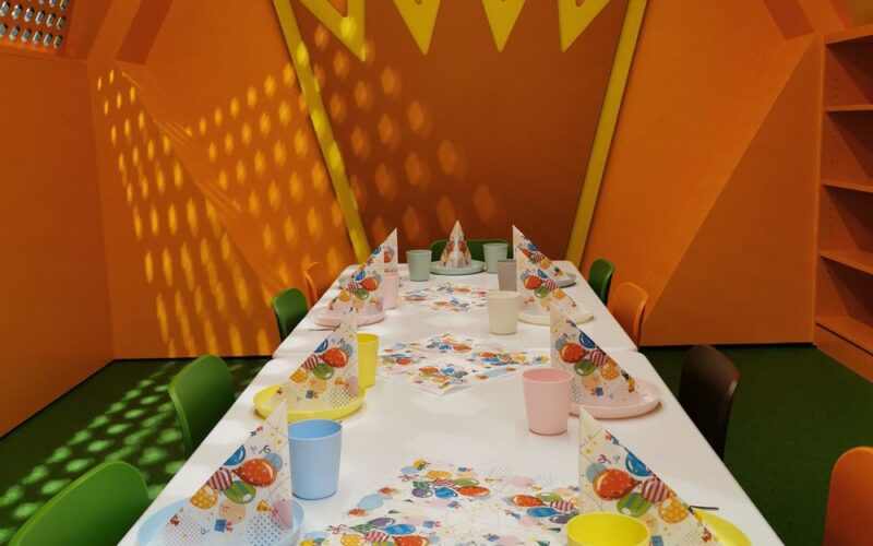 In einem Raum mit orangefarbenen Wänden und einer großen gelben Kronendekoration an der Rückwand ist ein bunter Partytisch mit Papierhüten, Bechern und Servietten gedeckt.