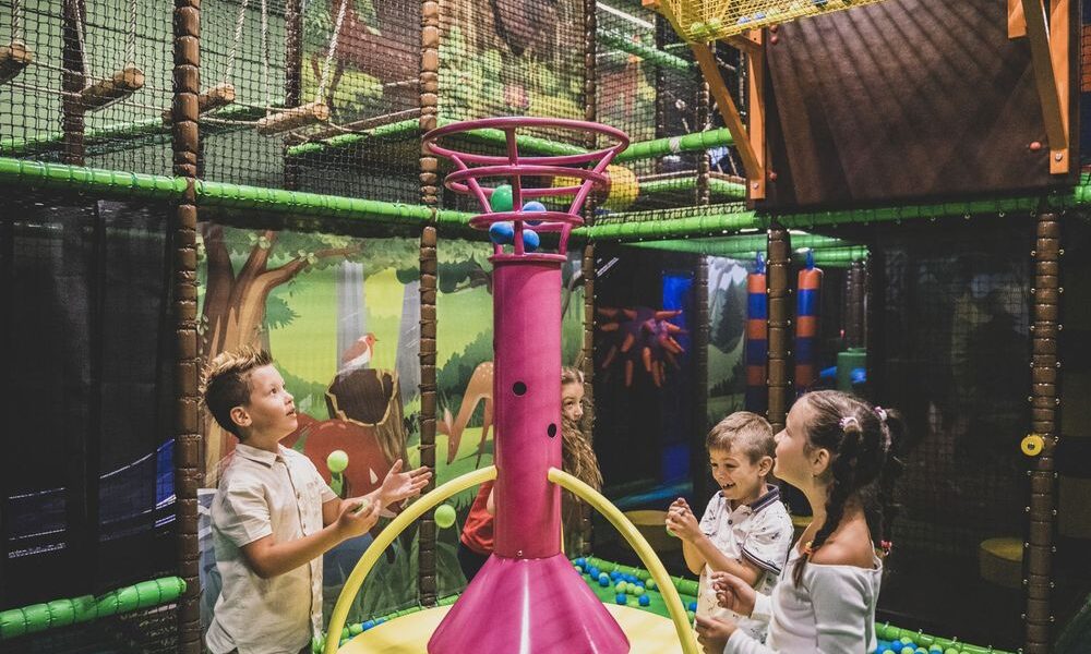 Vier Kinder spielen mit bunten Bällen um ein großes rosafarbenes Spielgerät in einem Innenspielbereich mit Dschungeldekoration.