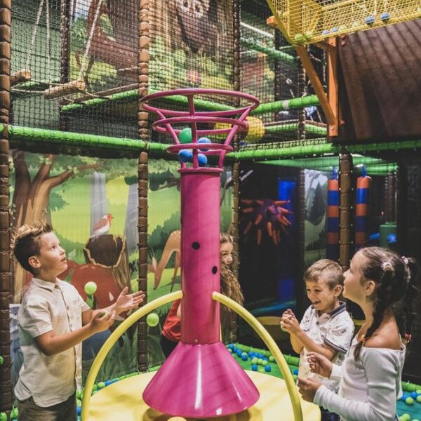 Vier Kinder spielen mit bunten Bällen um ein großes rosafarbenes Spielgerät in einem Innenspielbereich mit Dschungeldekoration.