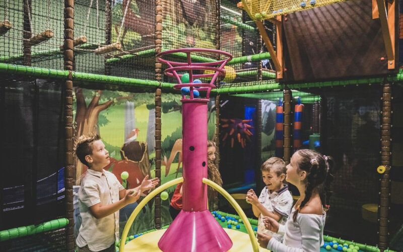 Vier Kinder spielen mit bunten Bällen um ein großes rosafarbenes Spielgerät in einem Innenspielbereich mit Dschungeldekoration.