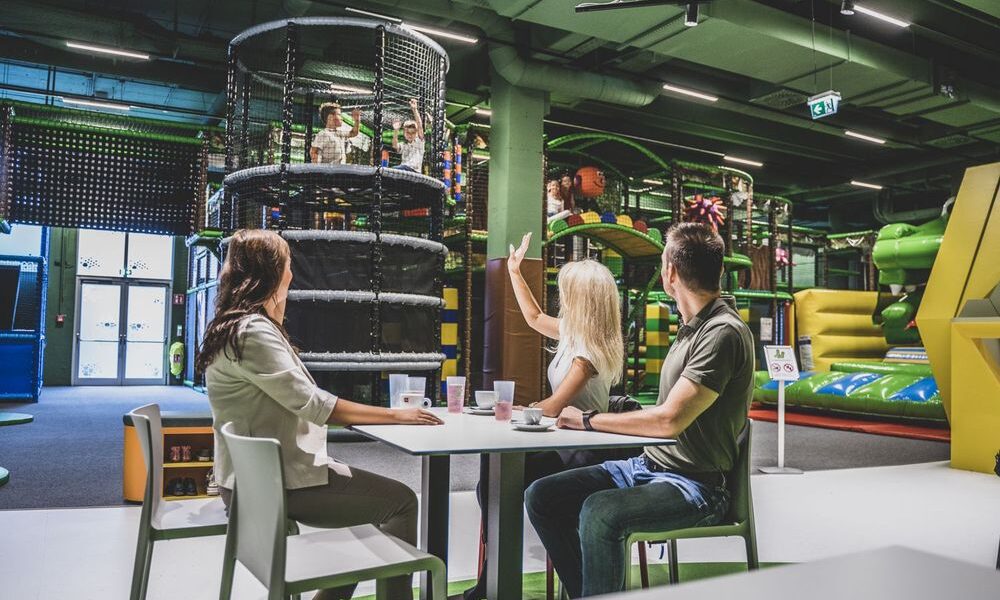 Drei Erwachsene sitzen an einem Tisch mit Getränken und schauen Kindern zu, die auf Indoor-Spielgeräten im Hintergrund spielen.