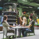 Drei Erwachsene sitzen an einem Tisch mit Getränken und schauen Kindern zu, die auf Indoor-Spielgeräten im Hintergrund spielen.