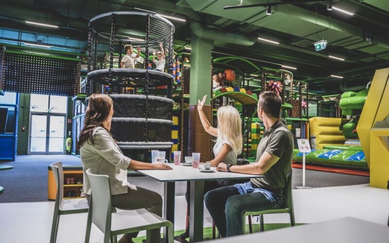 Drei Erwachsene sitzen an einem Tisch mit Getränken und schauen Kindern zu, die auf Indoor-Spielgeräten im Hintergrund spielen.