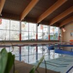 Hallenbad mit Bahntrennung und großen Fenstern, die den Blick auf eine verschneite Waldlandschaft freigeben; Sitzgelegenheiten am Beckenrand und Deckenbalken sind sichtbar.