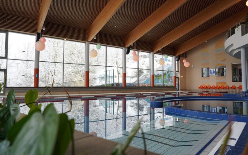 Hallenbad mit Bahntrennung und großen Fenstern, die den Blick auf eine verschneite Waldlandschaft freigeben; Sitzgelegenheiten am Beckenrand und Deckenbalken sind sichtbar.