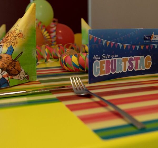 Ein für eine Geburtstagsfeier gedeckter Tisch mit bunten Servietten mit Cartoon-Tieren, gestreiften Tischsets, Luftballons und einer Karte mit der Aufschrift "Alles Gute zum Geburtstag".