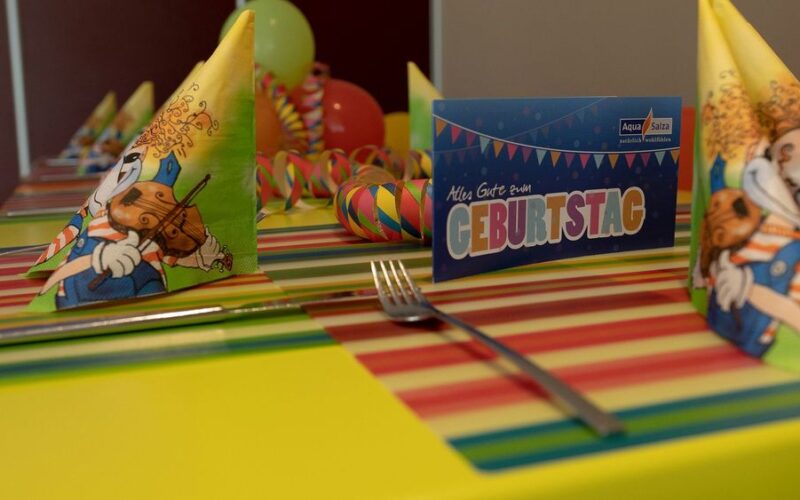 Ein für eine Geburtstagsfeier gedeckter Tisch mit bunten Servietten mit Cartoon-Tieren, gestreiften Tischsets, Luftballons und einer Karte mit der Aufschrift "Alles Gute zum Geburtstag".