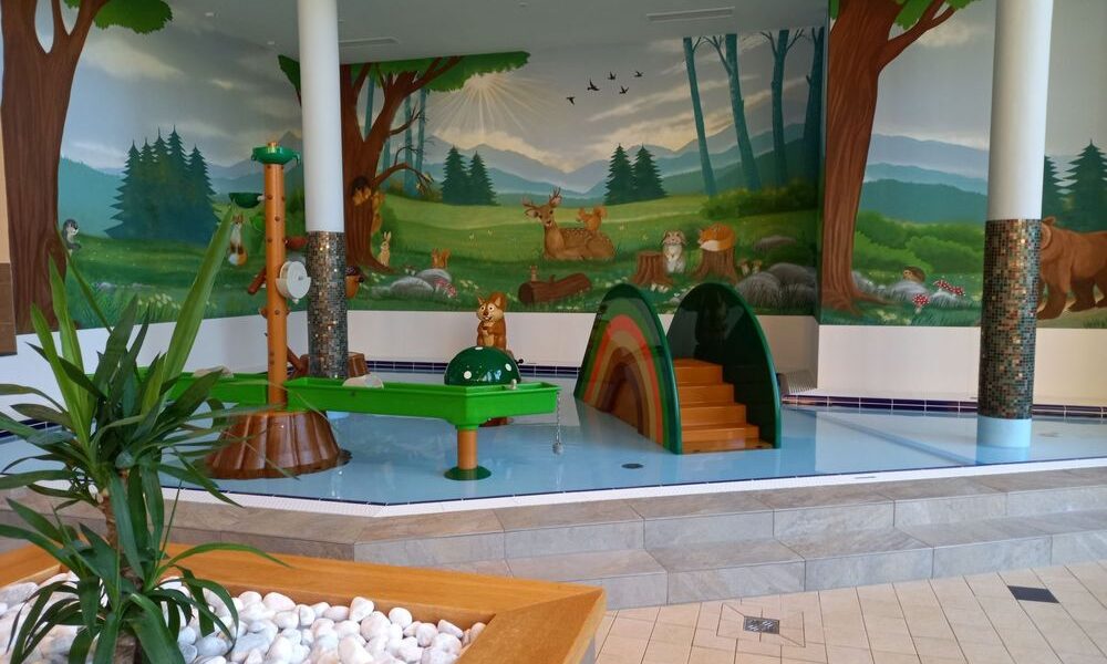 Kinderspielplatz im Innenbereich mit einem flachen Schwimmbecken, Spielgeräten mit Tiermotiven und einem Wandgemälde mit Waldtieren an den Wänden. Im Vordergrund befinden sich weiße Steine und eine Pflanze.