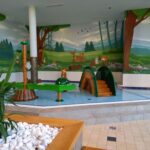 Kinderspielplatz im Innenbereich mit einem flachen Schwimmbecken, Spielgeräten mit Tiermotiven und einem Wandgemälde mit Waldtieren an den Wänden. Im Vordergrund befinden sich weiße Steine und eine Pflanze.